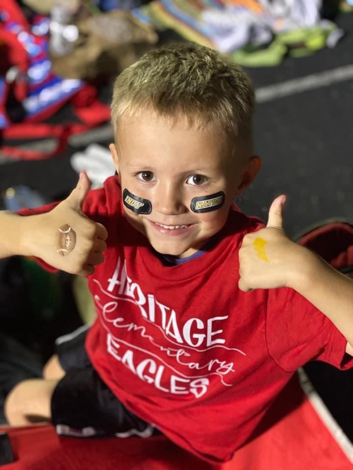 A young boy smiles while giving a thumbs at on the bleachers at a football game.